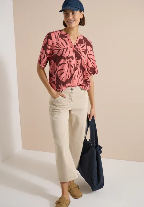 Model trägt rosa-schwarze Bluse mit Blattmuster, helle Culotte, beige Schuhe, blaue Tasche und Mütze.