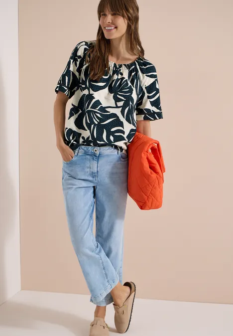 Carmen-Bluse mit Kurzarm und Blätterprint palm breeze green Frau trägt gemustertes Top, helle Jeans und beige Hausschuhe, hält eine orange Jacke in der Hand.
