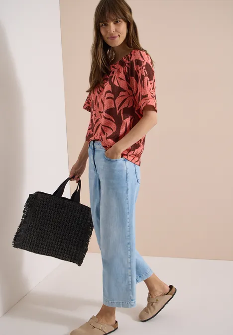 Carmen-Bluse mit Kurzarm und Blätterprint coral Frau in rotem Tropenprint-Oberteil, hellblauen Culotte-Jeans, beigen Slip-On-Schuhen und schwarzer Korbtasche.