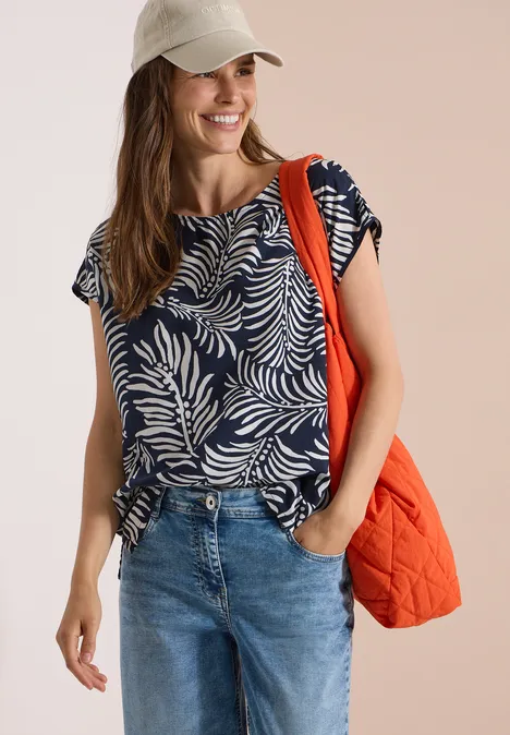 Dropped Shoulder Shirt mit Print universal blue Frau in Jeans, navyblauem Blattmuster-Shirt und Cap trägt eine orangefarbene gesteppte Tasche.