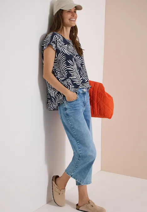 Dropped Shoulder Shirt mit Print universal blue Frau lehnt an Wand, trägt gemustertes Top, weite Jeans, bequeme Schuhe und hält eine orange Tasche.