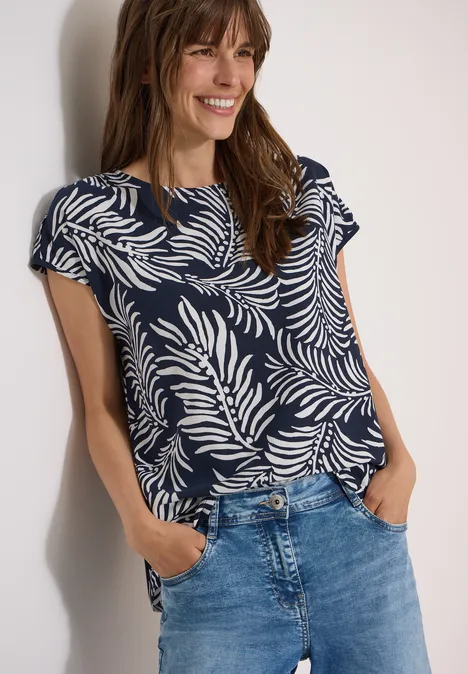 Dropped Shoulder Shirt mit Print universal blue Eine Person trägt ein dunkelblaues Shirt mit weißem Blattmuster und Jeans und hat die Hände in den Taschen.