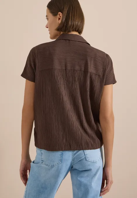 Frau von hinten in braunem Kurzarmhemd mit hellem Kragen und hellblauen Jeans.