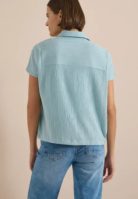 Eine Person von hinten trägt eine hellblaue kurzärmelige Bluse und Jeans vor beigem Hintergrund.
