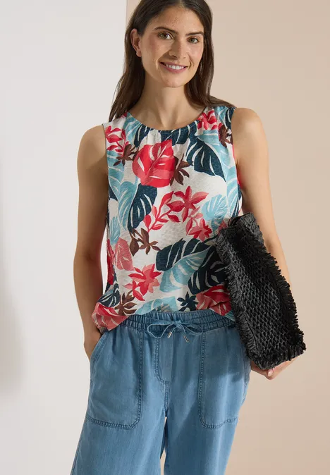 Vrouw draagt mouwloos shirt met rood bloemmotief, blauwe broek en houdt een zwarte tas vast.