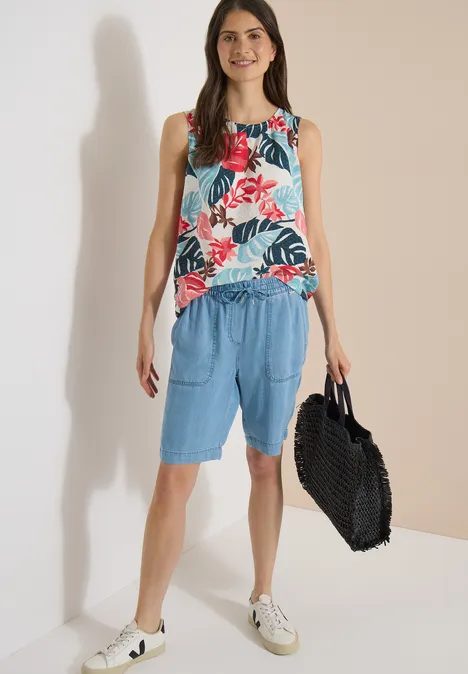 Vrouw in mouwloos bloemen-top, losse blauwe shorts, witte sneakers en grote zwarte tas.