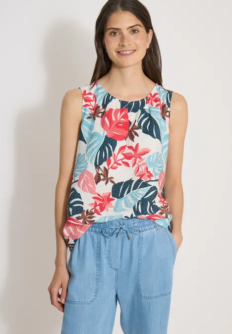 Vrouw draagt mouwloze, kleurrijke bloementop en blauwe broek, staat met handen in de zakken.
