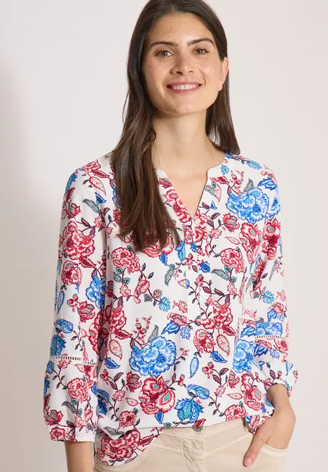 Frau trägt weiße Bluse mit blau-rotem Blumenmuster, V-Ausschnitt und langen Ärmeln.