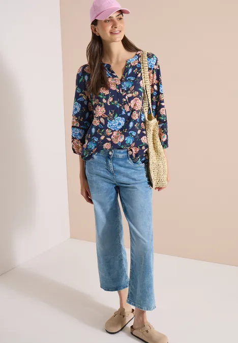 Eine Frau trägt eine lockere, blau florale Bluse, gerade Jeans, Basttasche und beige Loafers vor hellem Hintergrund.