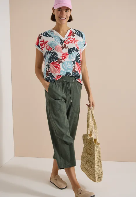 Frau trägt buntes, florales Top, weite grüne Hose und hält eine Strohtasche.