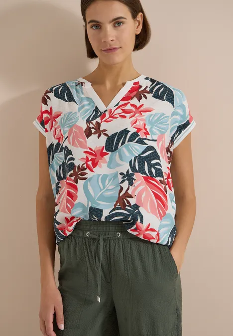 Person trägt bunte, floral gemusterte Bluse mit V-Ausschnitt und dunkelgrüner Hose mit Taschen.