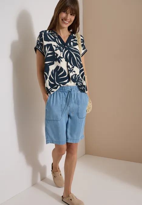 Frau trägt gemustertes kurzärmeliges Oberteil, lockere Jeans-Shorts und Espadrilles, dazu eine runde Strohtasche.
