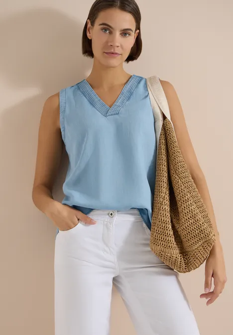 Frau in hellblauem ärmellosen V-Ausschnitt-Top, weißen Hosen und großer Strohtasche vor neutralem Hintergrund.