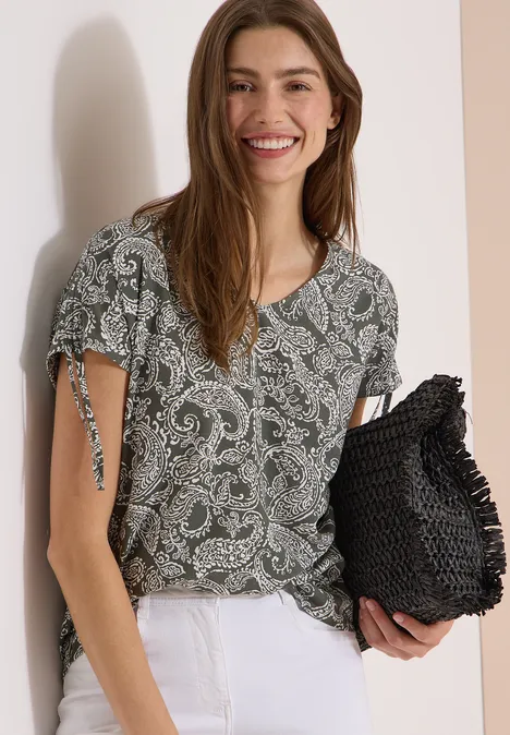 Overhemd met gerimpeld detail khaki Vrouw in een groen paisleyblouse met korte mouwen, witte broek en een zwarte gehaakte clutch.