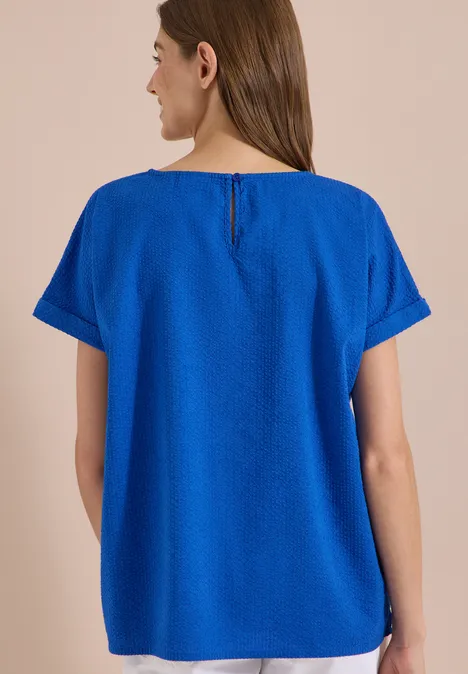 Blouse met ronde hals en knoopdetail surf blue Een vrouw van achteren draagt een los, felblauw shirt met korte mouwen en ronde hals.