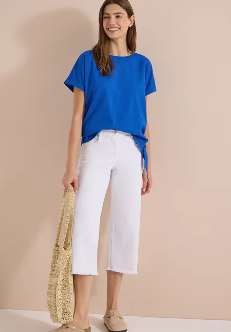 Blouse met ronde hals en knoopdetail surf blue Vrouw voor beige achtergrond met blauwe t-shirt, witte culottes, espadrilles en een gevlochten tas.