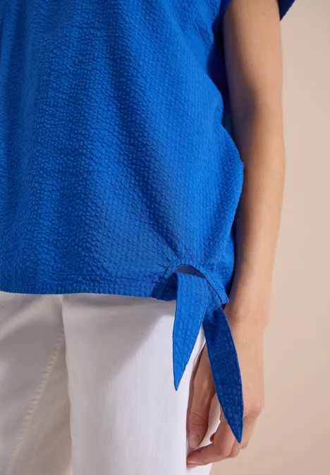Blouse met ronde hals en knoopdetail surf blue Close-up van persoon in blauw shirt met knoop aan zoom en witte broek.
