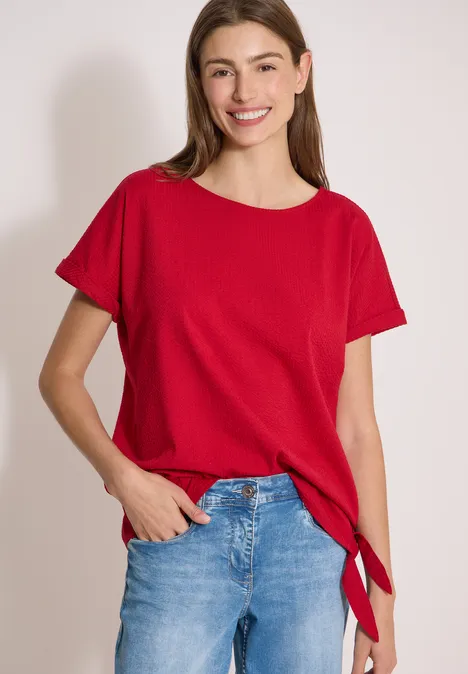 Blouse met ronde hals en knoopdetail tamarillo red Vrouw in rood T-shirt en jeans staat voor een witte achtergrond.