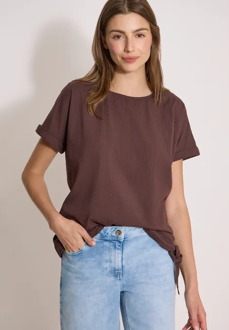 Blouse met ronde hals en knoopdetail brown cacao Vrouw draagt bruin T-shirt en lichte jeans tegen een neutrale achtergrond.