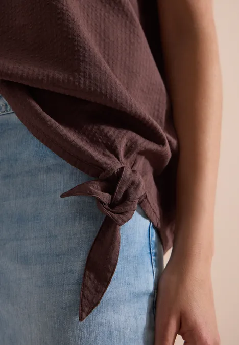 Blouse met ronde hals en knoopdetail brown cacao Close-up van een bruin topje met knoop aan de zijkant, gecombineerd met een lichtblauwe jeans en zichtbare arm.