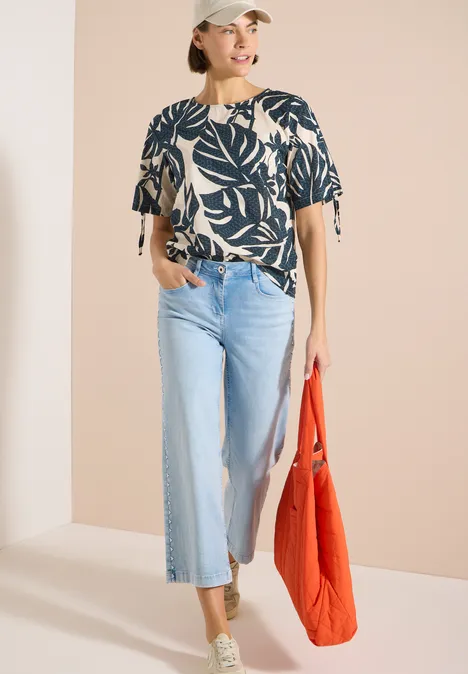 Blouse avec col rond et fronces palm breeze green Femme en chemisier à motif de feuilles, jean large clair, baskets vertes et sac rouge.