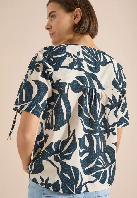 Blouse avec col rond et fronces palm breeze green Femme porte un chemisier blanc à motif de feuilles bleu foncé, manches mi-longues et liens sur les côtés.