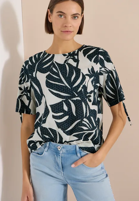 Blouse avec col rond et fronces palm breeze green Femme en T-shirt blanc à manches courtes imprimé de feuilles vert foncé, rentré dans un jean bleu clair devant fond beige.