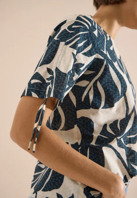 Blouse avec col rond et fronces palm breeze green Gros plan d'une femme portant un haut blanc à motifs de feuilles bleues, manches courtes à nouer, et pantalon clair.