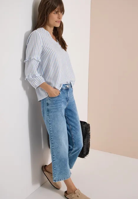 Frau lehnt an Wand, trägt gestreifte Bluse, lockere cropped Jeans und flache Loafers, hält Tasche.