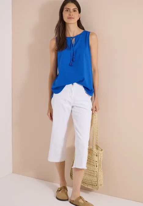 Frau mit blauem Top, weißen Culotte-Hosen und Strohtasche, trägt Sandalen.