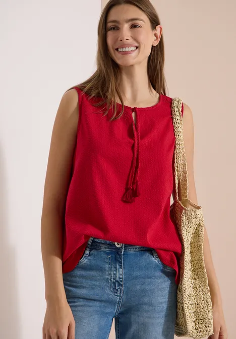 Blusentop mit Split Neck und Bändern tamarillo red Frau in rotem ärmellosem Oberteil, Jeans und Beuteltasche mit Muster.