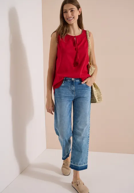 Blusentop mit Split Neck und Bändern tamarillo red Frau in rotem ärmellosem Top, weiten blauen Jeans und geflochtener Strohtasche vor heller Wand.