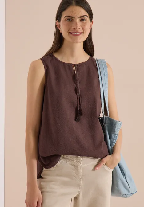 Frau trägt ärmelloses, braunes Top mit Quasten, helle Hose und blaue Jeans-Tasche über der Schulter.
