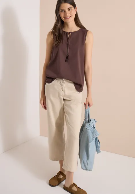 Frau in braunem Top, heller Culotte-Hose und braunen Schuhen, hält hellblaue Jeansjacke.