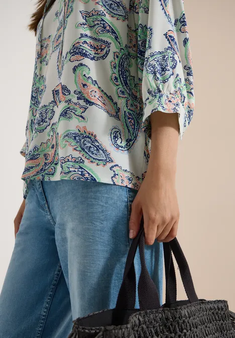 Person in gemusterter Bluse und Jeans hält eine schwarze geflochtene Korbtasche.