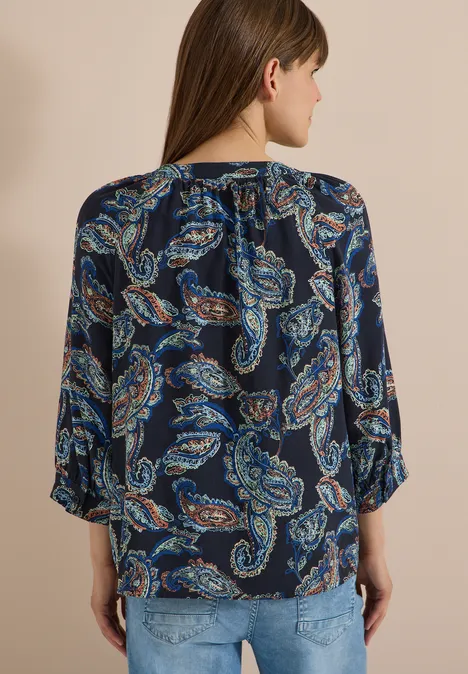 Frau von hinten in dunkelblauer Bluse mit Paisley-Print und Jeans vor beigem Hintergrund.