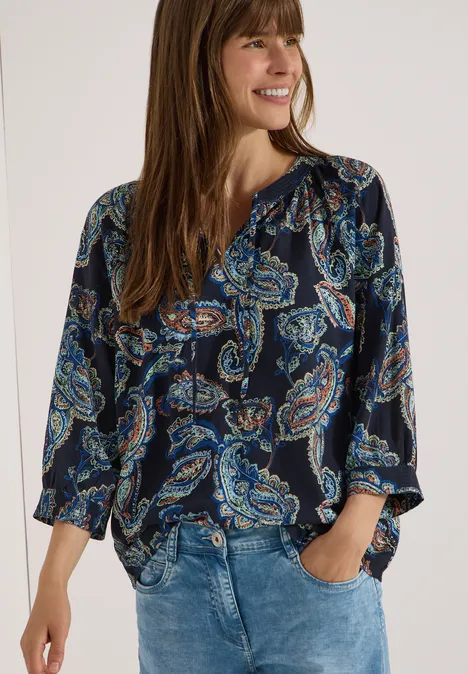 Frau in Bluse mit Paisley-Print und Jeans, Hände in den Taschen.