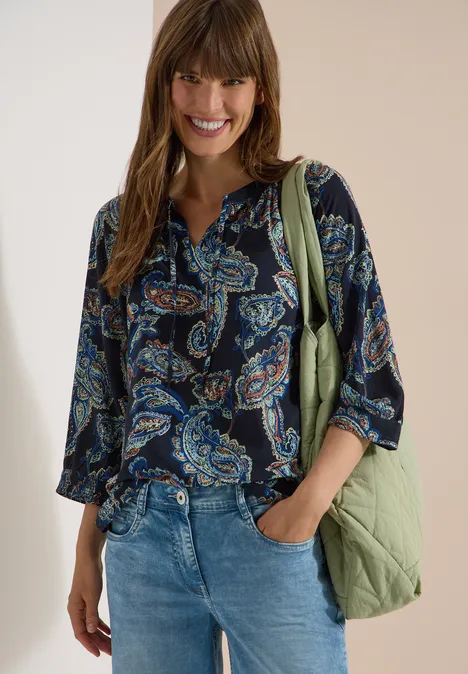 Frau in dunkler Paisley-Bluse, Jeans und grüner Tasche, entspannt stehend.