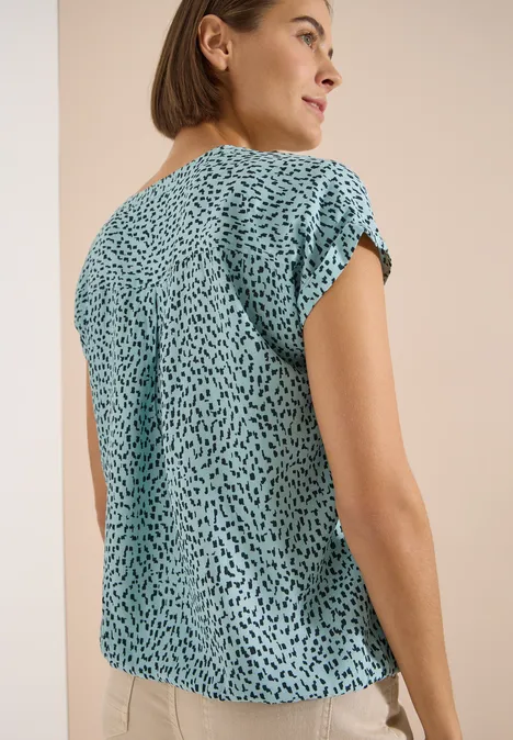 Rückenansicht einer Frau in einem türkisfarbenen, kurzärmeligen Shirt mit schwarzem Tupfenmuster.