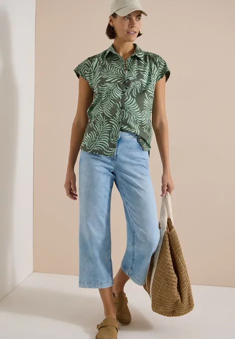 Frau mit grüner, gemusterter Kurzarmbluse, Cropped-Jeans, beigen Mules und Strohtasche vor hellem Hintergrund.