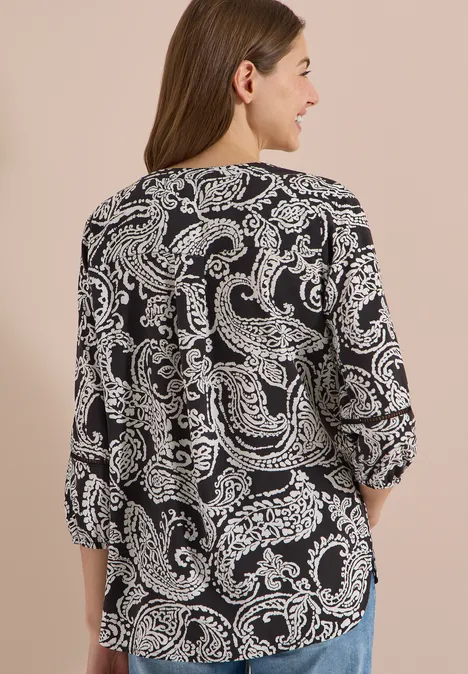 Tuniek met 3/4-mouwen en print Black Vrouw van achteren in een zwarte blouse met wit paisleypatroon voor een beige achtergrond.