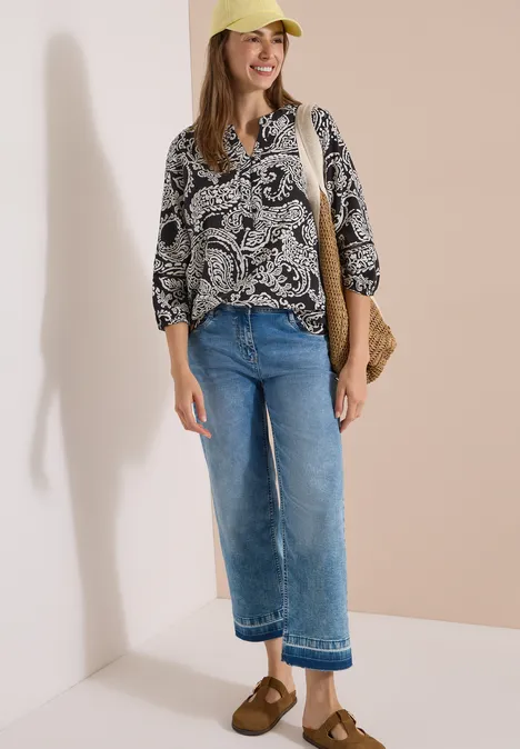 Tuniek met 3/4-mouwen en print Black Vrouw draagt gebloemde blouse, blauwe jeans, bruine sandalen, strooien tas en gele hoed tegen lichte achtergrond.