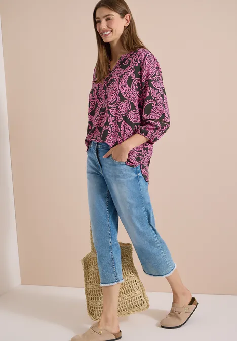 Tuniek met 3/4-mouwen en print raw khaki Een vrouw in een roze top met bladprint, blauwe jeans, beige mules en een strooien tas.