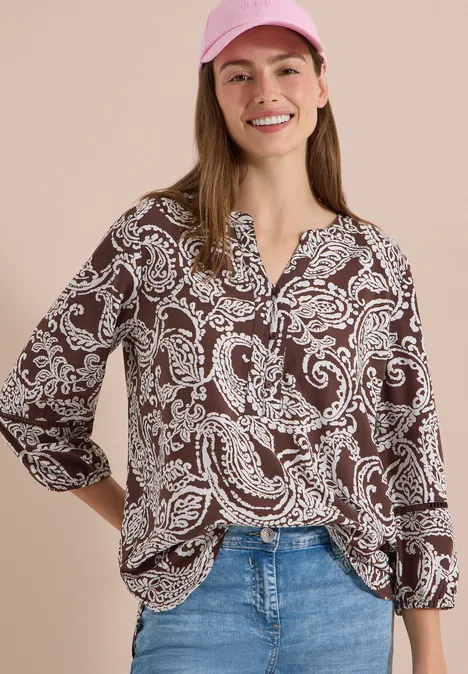 Tuniek met 3/4-mouwen en print brown cacao Vrouw met bruine blouse met wit paisleymotief, spijkerbroek en roze pet voor lichte achtergrond.