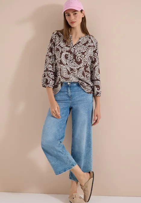 Tuniek met 3/4-mouwen en print brown cacao Vrouw in paisleyblouse, wijde blauwe jeans en lichte espadrilles voor een egale achtergrond.