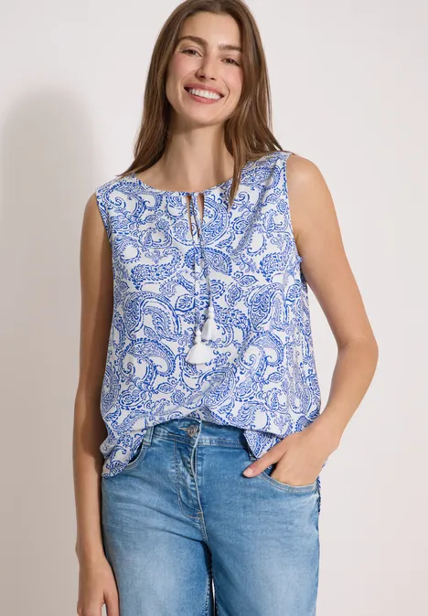 Frau in blau-weißem Paisley-Top mit Quasten und Jeans, vor neutralem Hintergrund.