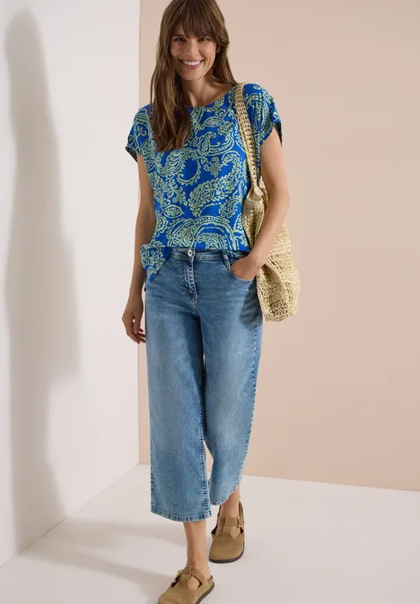 Blusenshirt mit Rundhals und Print surf blue Frau trägt blau gemustertes T-Shirt, helle Culotte-Jeans, Strohtasche und beige Keilsandalen.
