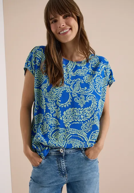 Blusenshirt mit Rundhals und Print surf blue Frau trägt blauen, gemusterten Kurzarmbluse und Jeans vor hellem Hintergrund.