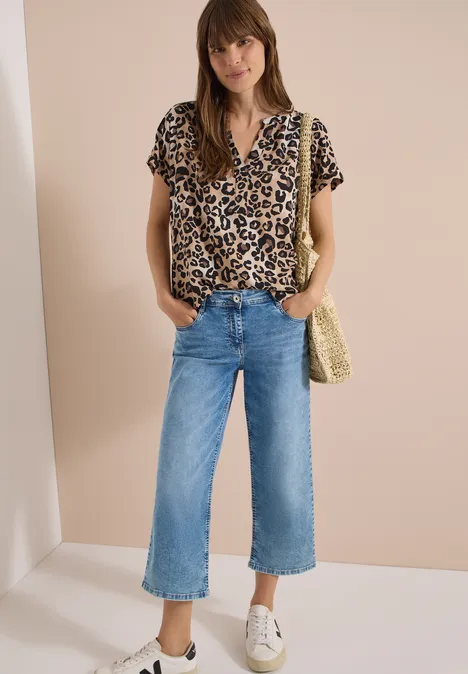 Frau mit Leopardenbluse, weiten Jeans-Culottes, weißen Sneakern und Strohtasche vor beiger Wand.