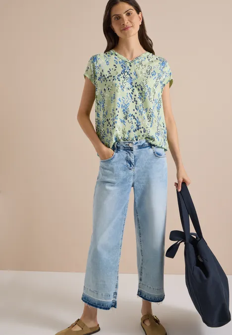 Chemisier à manches courtes avec mélange de textures raw matcha lime Eine Frau trägt eine geblümte Bluse, weite helle Jeans und hält eine dunkelblaue Tasche.